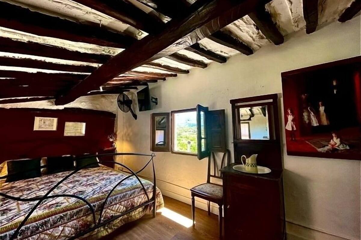 A rustic room with a bed, a chair, a small table, and a mirror. There are two windows and a view of the outdoors.