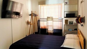 Un dormitorio con cama, televisor, estante con objetos y ventana.
