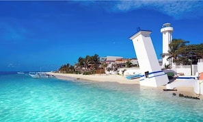 Une plage à l’eau turquoise et limpide, un bâtiment blanc surmonté d’un phare, et des bateaux visibles au loin.