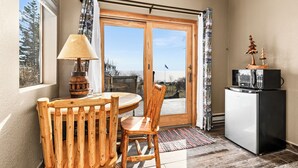 Une petite pièce avec une table et des chaises en bois, une lampe, un réfrigérateur et une vue sur l’extérieur, visible à travers une porte vitrée.
