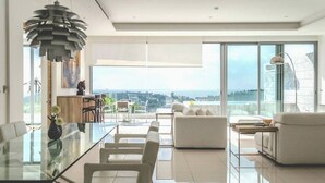 A modern living room with a glass dining table, white sofas, and a view of the cityscape through large windows.
