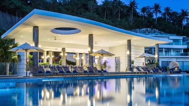 A modern poolside area with lounge chairs and umbrellas, a clear pool, and a building with a flat roof and large pillars.