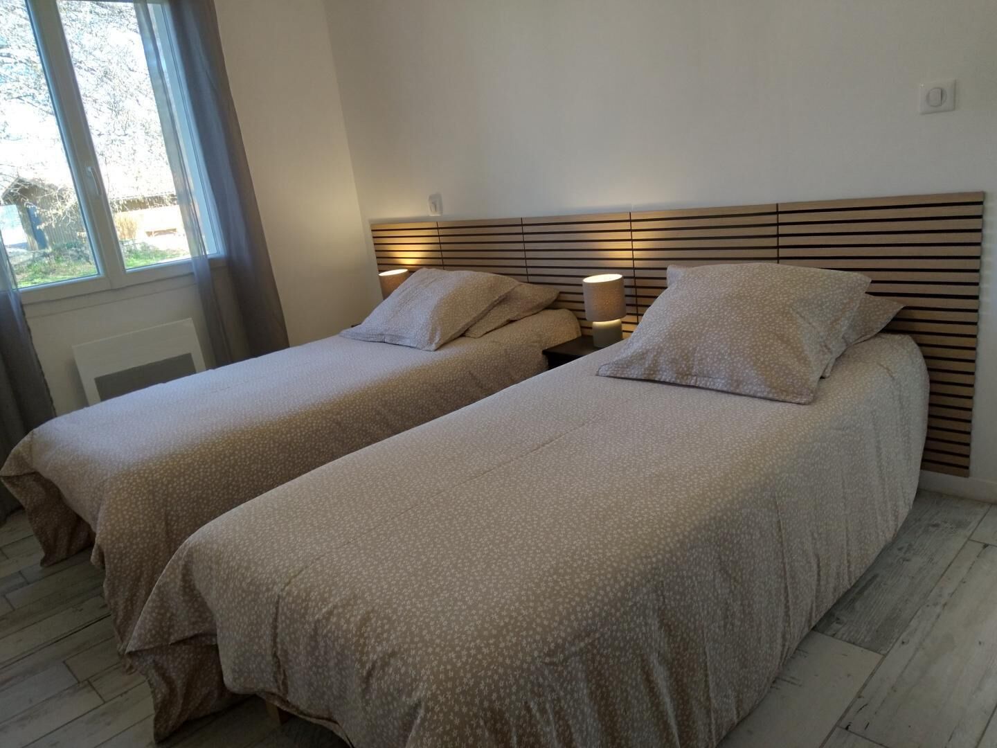 Een slaapkamer met twee eenpersoonsbedden, een houten hoofdbord, nachtlampjes en een raam met gordijnen.