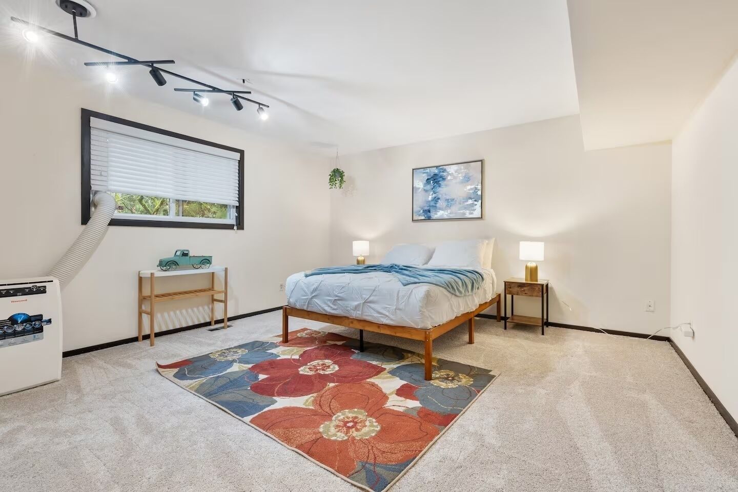 Un dormitorio con cama, mesitas de noche, radiador, escritorio, silla, ventana con persianas y lámpara de techo, además de un cuadro colgado en la pared.