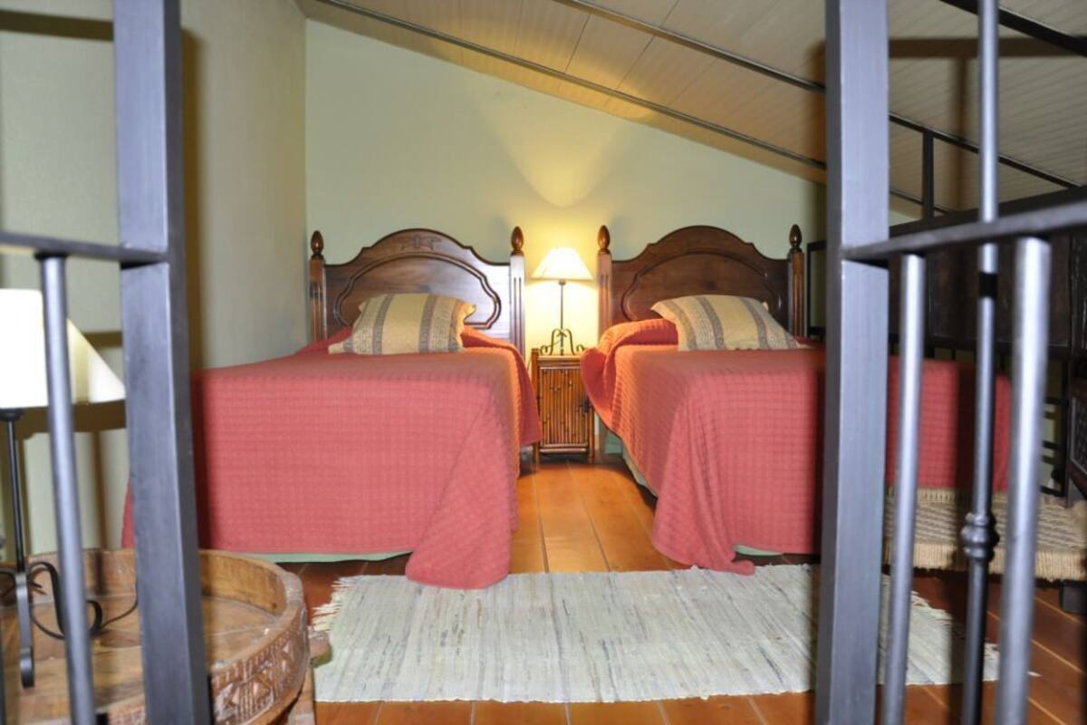 Een kamer met twee bedden, elk met een houten hoofdbord en rode beddengoed. Tussen de bedden staat een nachtkastje met een lamp, en in de hoek een klein tafeltje met een lamp. De vloer is betegeld en er ligt een wit vloerkleed.