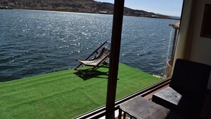 A view of a lake with a wooden chair on a grassy area, a wooden deck, and a building with a window. - INTI TITIKAKA (Puno)