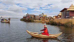 A person in a traditional boat on a calm river, surrounded by thatched-roof huts and a clear sky. - INTI TITIKAKA (Puno)