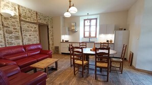 A cozy living room with a red leather sofa, a wooden coffee table, and a dining area with wooden chairs and a table. A stone wall and a window with a view are visible in the background. - Gîtes de France® - Au Refuge des Artistes (VIAZAC)