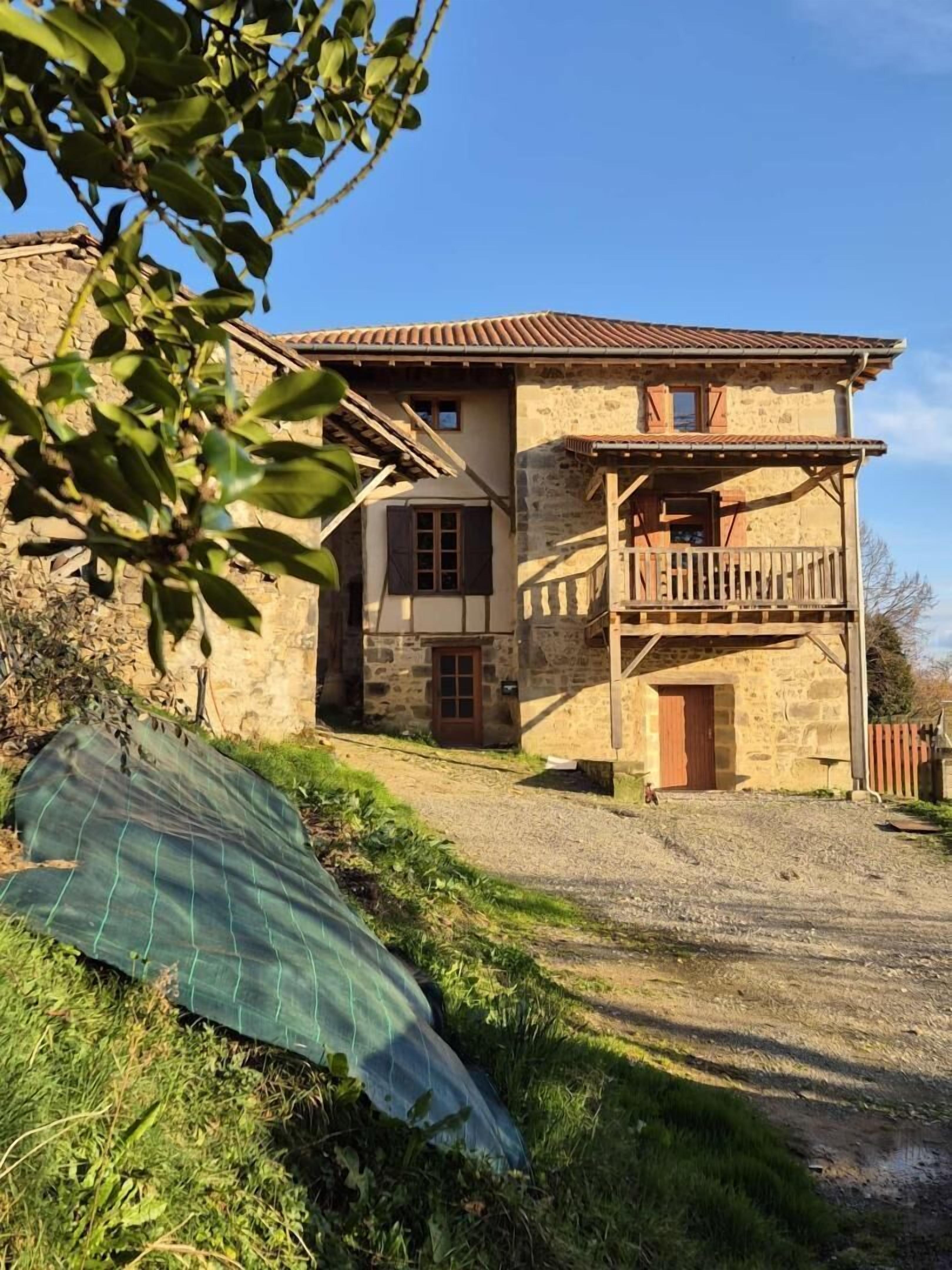A two-story stone house with a wooden balcony, a gravel driveway, and a green tarp on the ground.