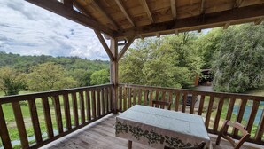 A wooden deck with a table and chairs, overlooking a lush green landscape. - Gîtes de France® - Au Refuge des Artistes (VIAZAC)