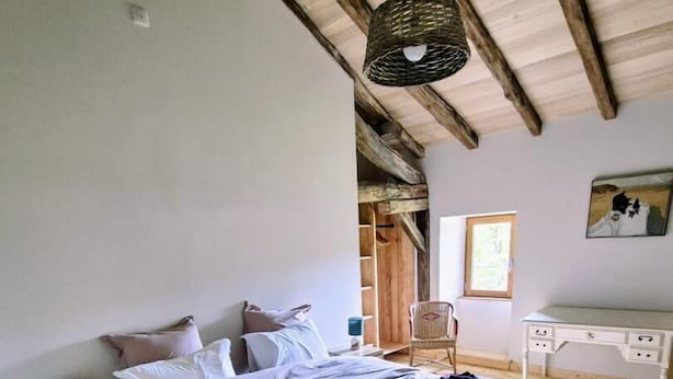 A bedroom with a wooden floor, a bed with a white duvet, a bedside table with a lamp, a wicker hanging light fixture, and a desk with a chair. - Gîtes de France® - Au Refuge des Artistes (VIAZAC)
