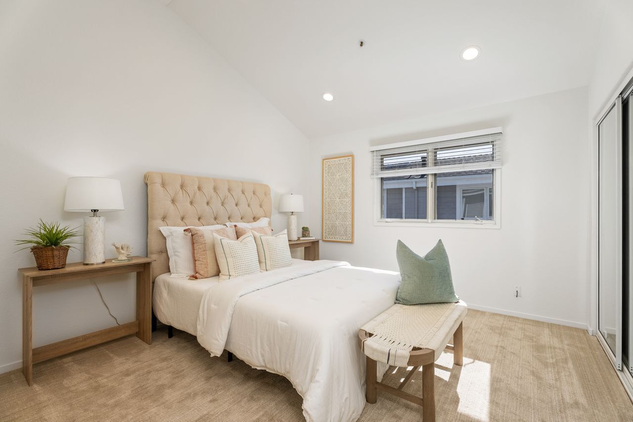 Une chambre à coucher avec une tête de lit capitonnée, un grand lit, une table de chevet avec une lampe, une petite table avec une plante et une fenêtre avec des stores.