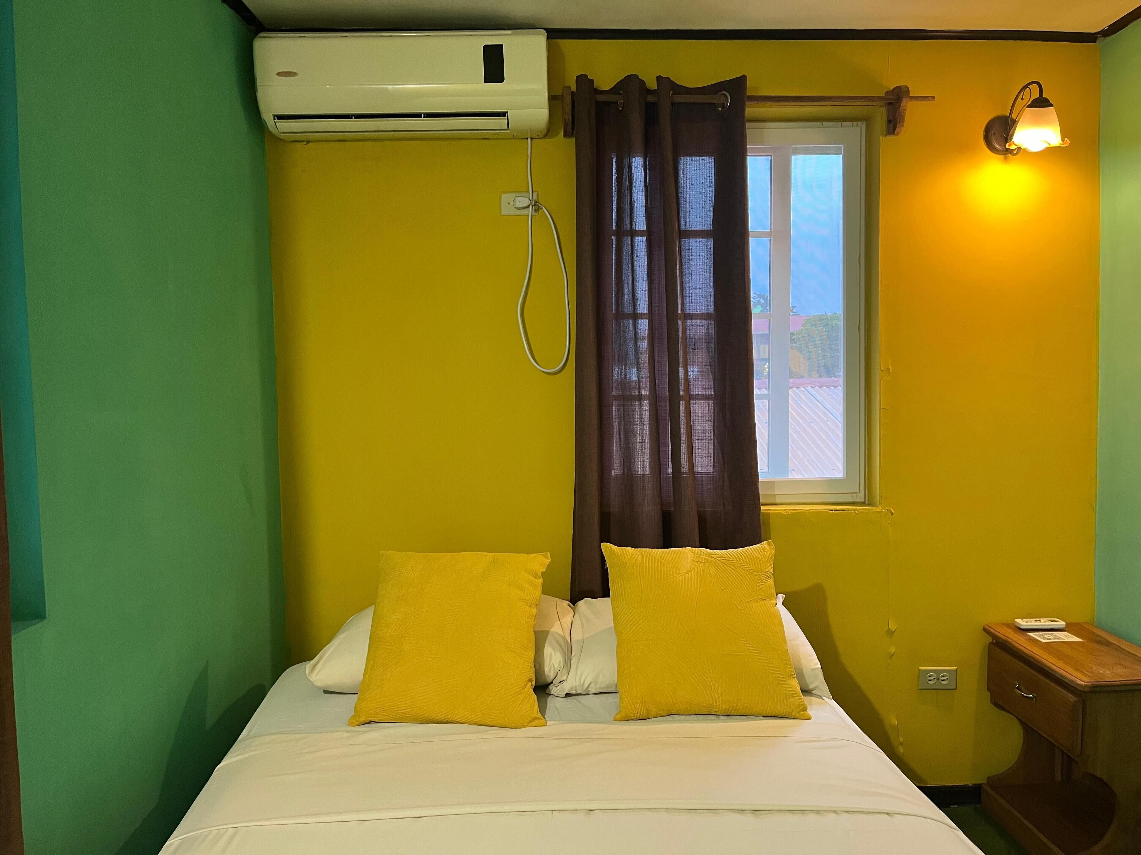 A room with a bed, a window with curtains, an air conditioning unit, and a bedside table with a lamp.