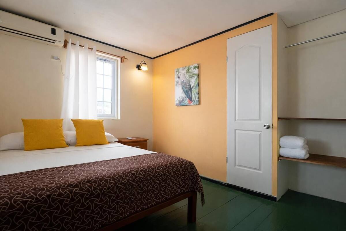A hotel room with a bed, a nightstand, a window with curtains, a door, and a wall-mounted shelf with towels.