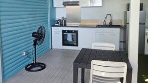 A compact kitchen with white cabinets, a built-in oven, and a sink. There's a dining table with two chairs and a fan on the floor.