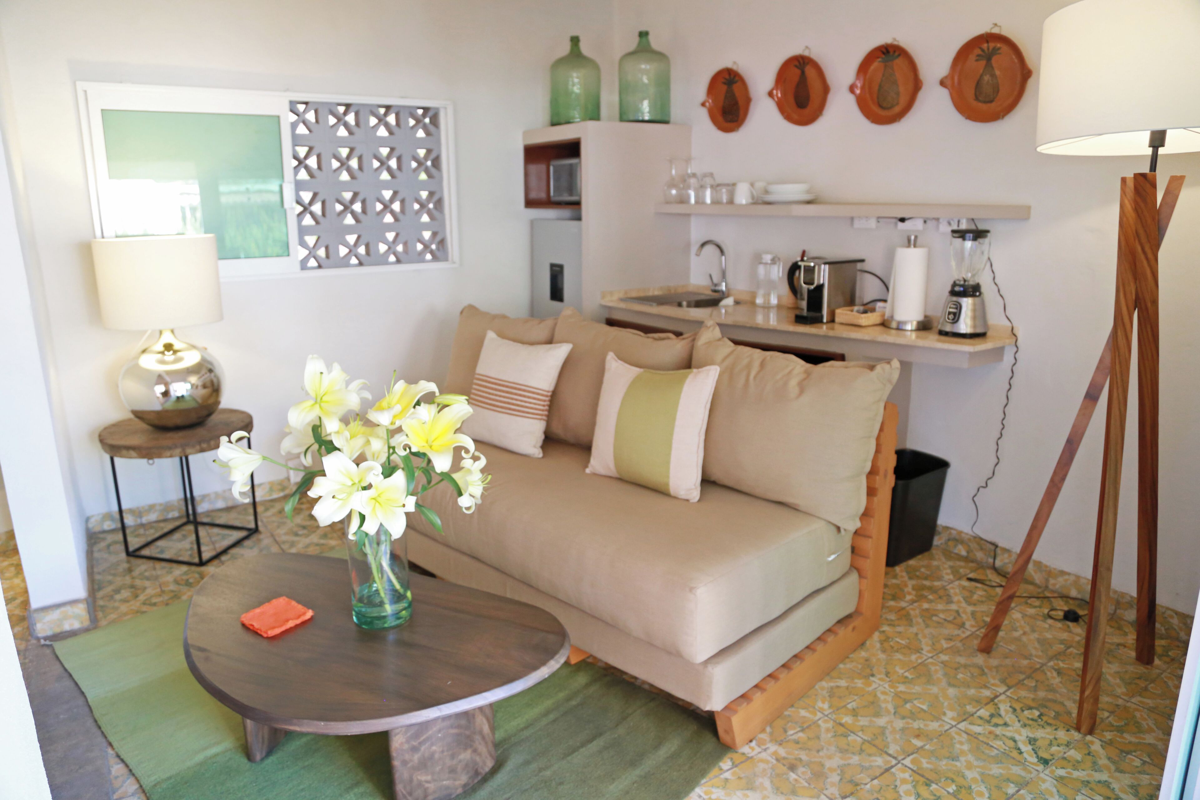 A living room with a sofa, a coffee table with a vase of flowers, a lamp, and a kitchen area with shelves and appliances.