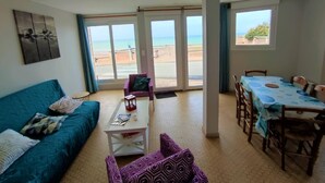 Een woonkamer met een bank, een salontafel en een eethoek met een tafel en stoelen. Er is een groot raam met uitzicht op het strand.