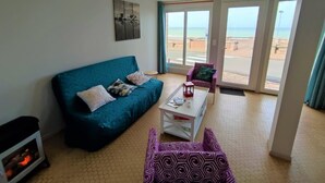 Een woonkamer met een blauwgroene bank, gekleurde kussens, een witte salontafel en een paarse fauteuil. Links is een open haard te zien, en de kamer heeft grote glazen deuren met uitzicht op een strand.