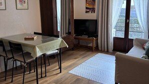 Una sala de estar con una mesa de comedor, sillas, un sofá, un televisor y un amplio ventanal con cortinas.