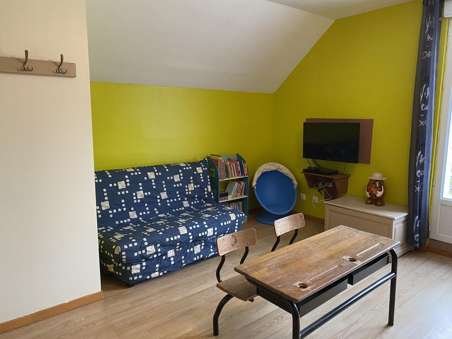 A room with a blue patterned sofa, a wooden table, a bookshelf, and a television on a wall.