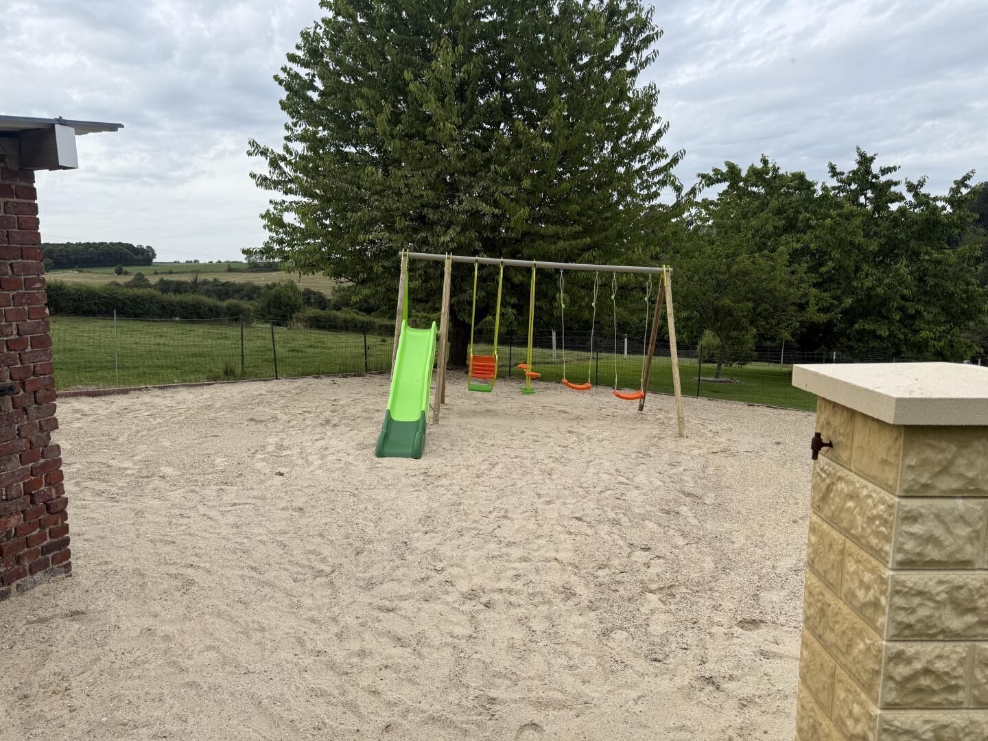 A playground with a green slide and swings, surrounded by a sandy area and a brick wall.