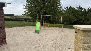 A playground with a green slide and swings, surrounded by a sandy area and a brick wall.