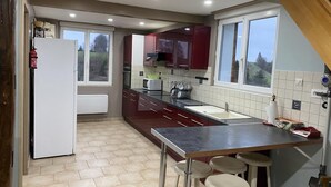 A compact kitchen with a refrigerator, stove, microwave, and a sink. There are bar stools and a window providing natural light. - Gîtes de France® - l'Humesnil (ST VICTOR L'ABBAYE)