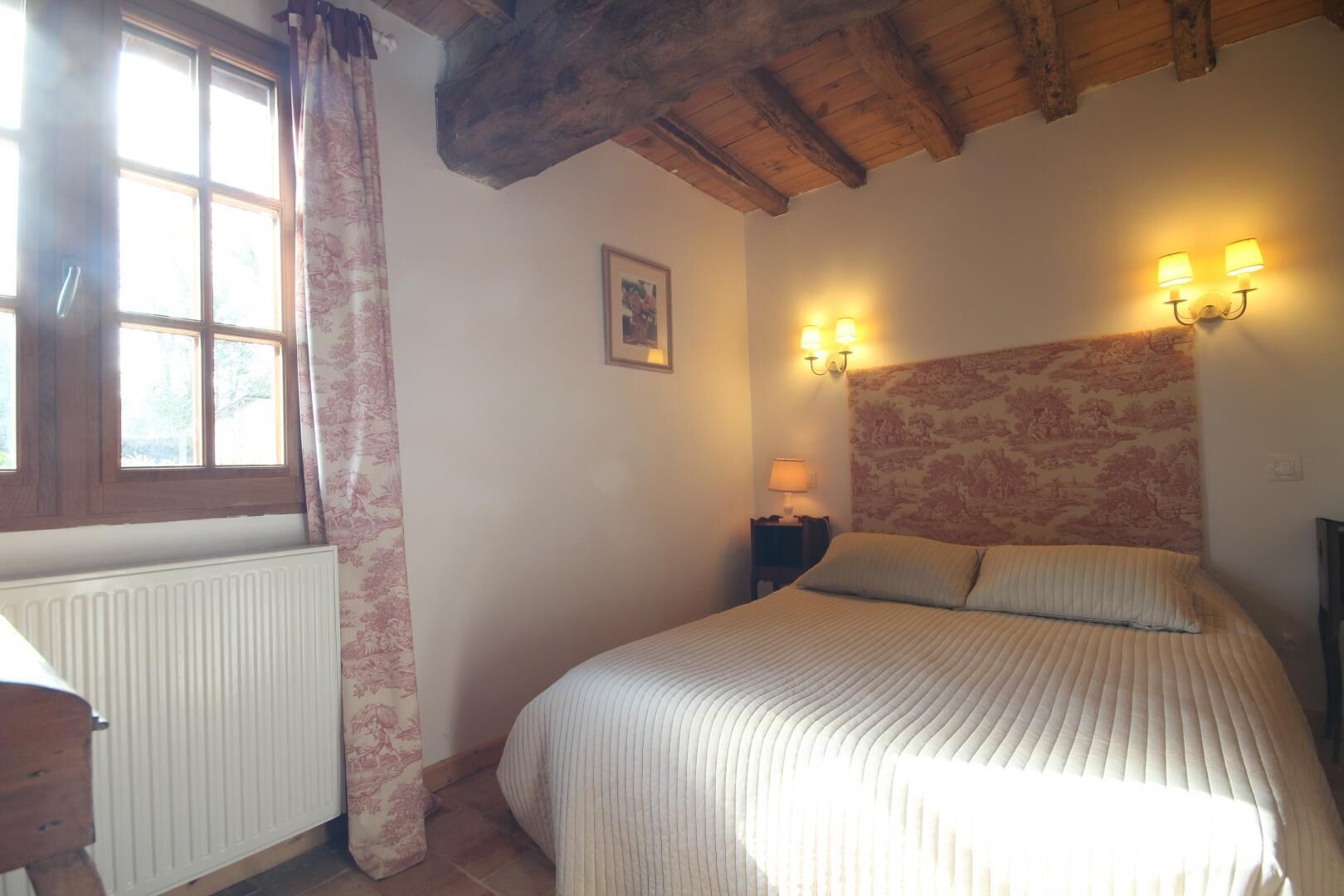Camera da letto con un letto, un comodino, una lampada, una finestra con tende e un soffitto in legno.