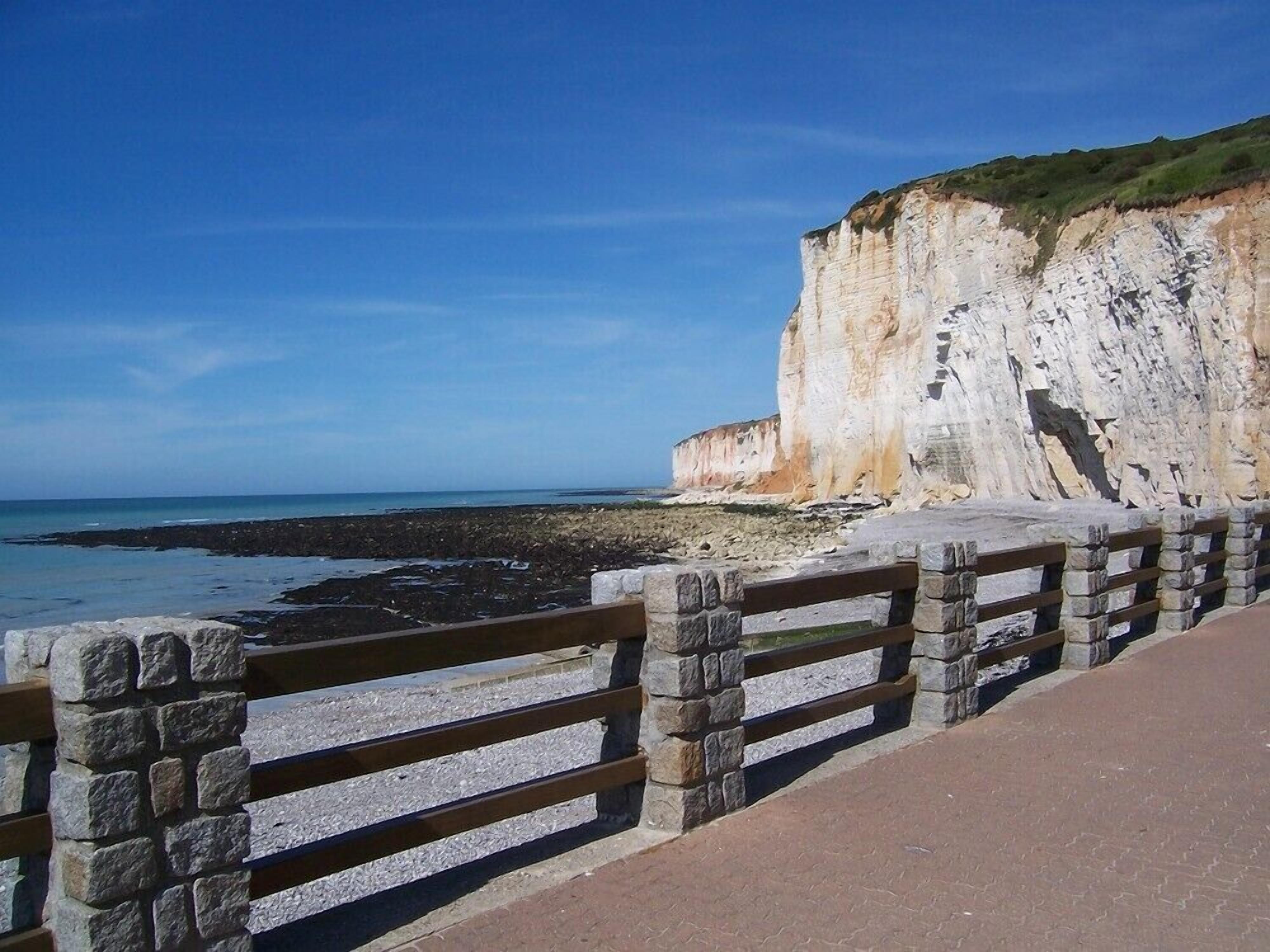 A coastal walkway with a stone railing, leading to a cliff with white rock formations and a view of the sea.
