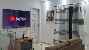 Un salón moderno con un televisor de pantalla plana mostrando YouTube, un sofá marrón, una mesa de centro de vidrio, un jarrón rojo y un cuadro en la pared.