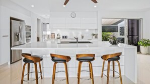 A modern kitchen with a central island, stainless steel appliances, wooden stools, and a ceiling fan.