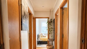 A hallway with wooden doors, a hanging plant, a bed with a window, and a small table with baskets. - The Coasty Bach | Cosy Cottage | Rest & Reset (Tolaga Bay)