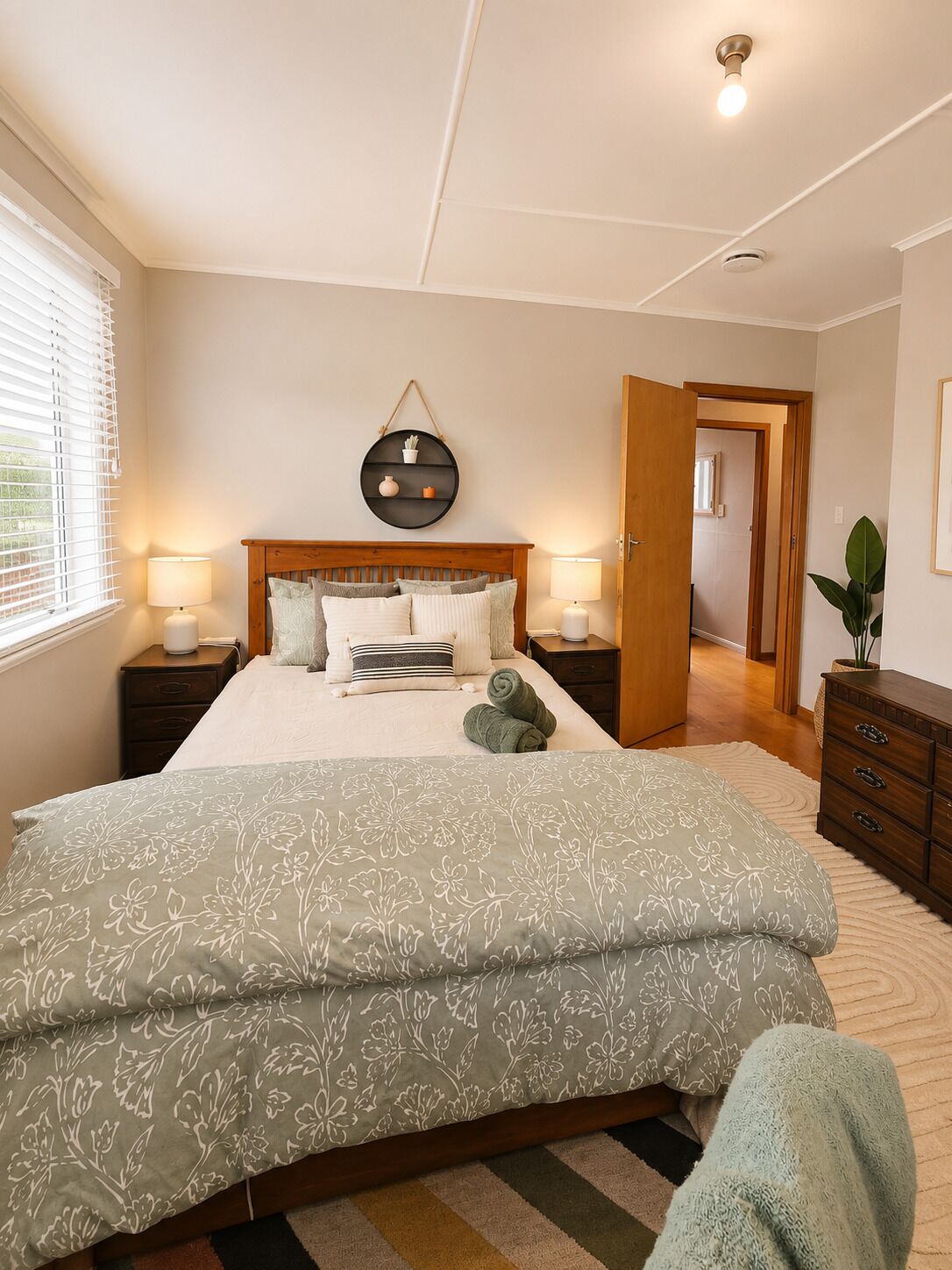 A bedroom with a bed, bedside tables, a wall-mounted shelf, a door leading to another room, and a window with blinds.