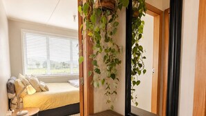A bedroom with a bed, bedside table, lamp, and a hanging plant. - The Coasty Bach | Cosy Cottage | Rest & Reset (Tolaga Bay)