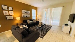 A living room with a sofa, a coffee table, and a TV. There are framed pictures on the wall and a window with curtains. - Bella Vida Home (Kissimmee)