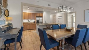 A modern kitchen with wooden cabinets, a central island, and a dining area with a wooden table and blue chairs. - Spacious 3BR Condo | Walk to Beach | Pool, Hot Tub & Balcony Views (Blue Mountain Beach)