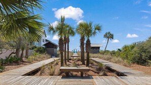 A wooden boardwalk leading to a beach area with palm trees and a building in the background. - Spacious 3BR Condo | Walk to Beach | Pool, Hot Tub & Balcony Views (Blue Mountain Beach)
