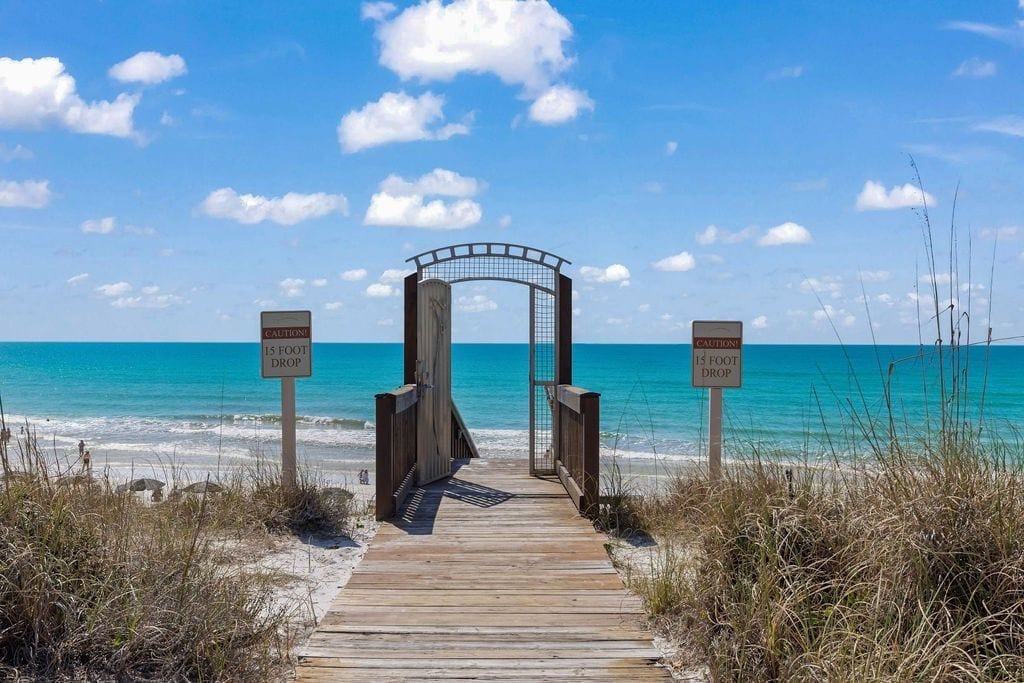 A wooden boardwalk leading to a beach with caution signs indicating a 15-foot drop.