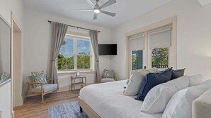 A bedroom with a bed, a ceiling fan, a TV, a window with curtains, and a view of greenery outside. - Spacious 3BR Condo | Walk to Beach | Pool, Hot Tub & Balcony Views (Blue Mountain Beach)