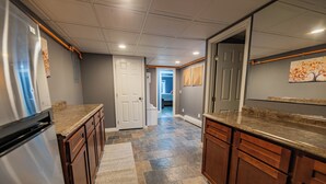 Una cocina con refrigerador, armarios de madera, piso de baldosas de piedra y una puerta que da a otra habitación.