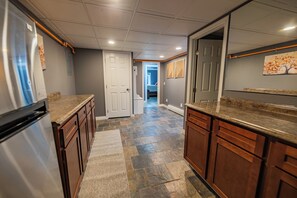 A kitchen with a refrigerator, wooden cabinets, a stone tile floor, and a door leading to another room.