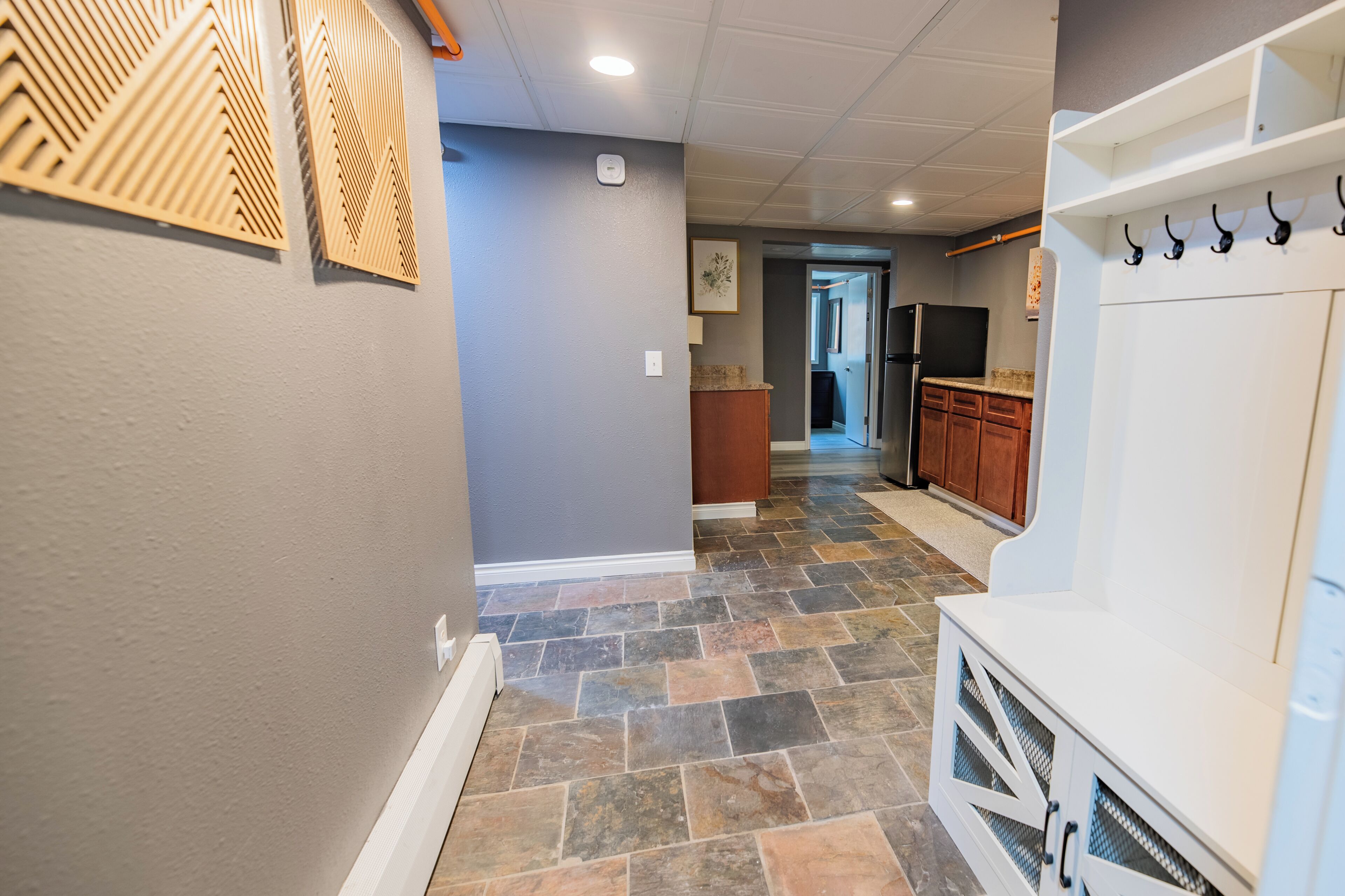 A hallway with a stone floor, a white bench, and a kitchen area with wooden cabinets and a refrigerator.