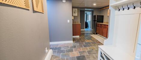 A hallway with a stone floor, a white bench, and a kitchen area with wooden cabinets and a refrigerator.