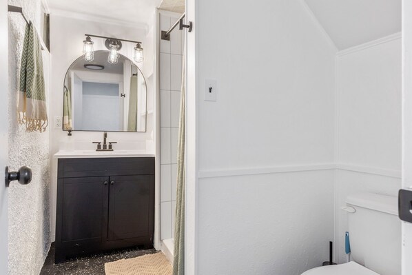 A bathroom with a white sink, dark cabinets, a mirror with lights, and a shower area.