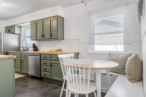 A kitchen with green cabinets, a white sink, and a wooden countertop. There is a white chair and a bench with cushions.