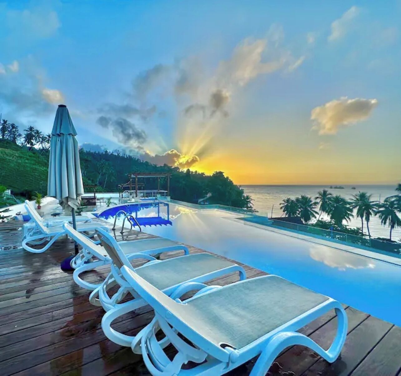 A poolside area with lounge chairs, a wooden deck, and a view of the ocean at sunset.