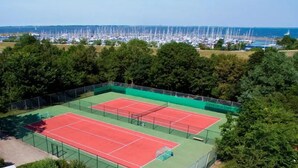 A tennis court with a net, surrounded by trees and a view of a marina with boats. - Chalet near the beach in Zeeland - perfect for water sports, cycling & relaxation (Brouwershaven)