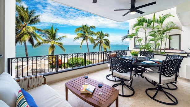 A balcony with a view of the sea, palm trees, and a dining area with chairs and a table. - Oceanfront Paradise | Pool & Walk-Out Beach Access (Puerto Vallarta)