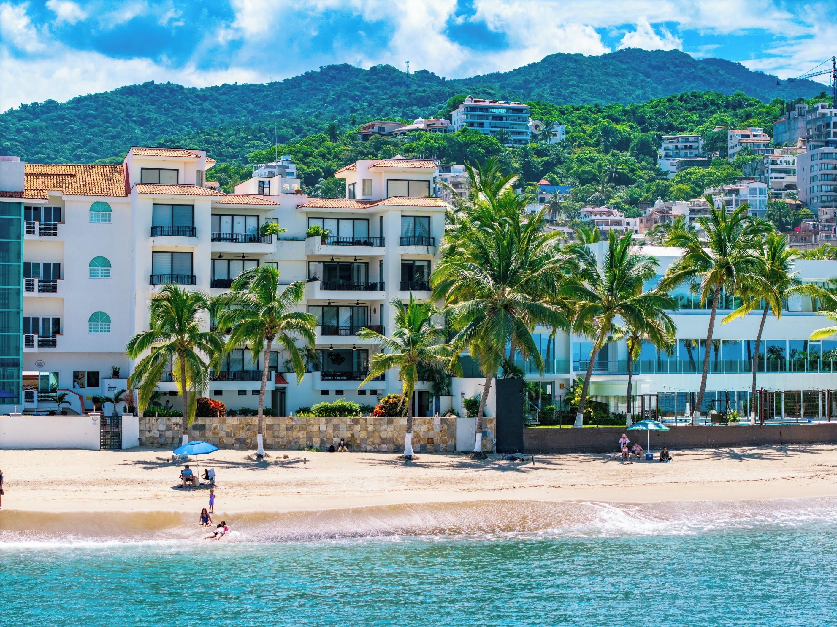 Un hôtel situé en bord de mer, doté de nombreux balcons, de palmiers et offrant une vue sur la plage et les montagnes.