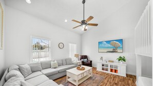 A spacious living room with a large sectional sofa, a coffee table, a ceiling fan, a painting of a beach, and a white cabinet with shelves. - "Casa Verde" - Private Pool - West End (Panama City Beach)